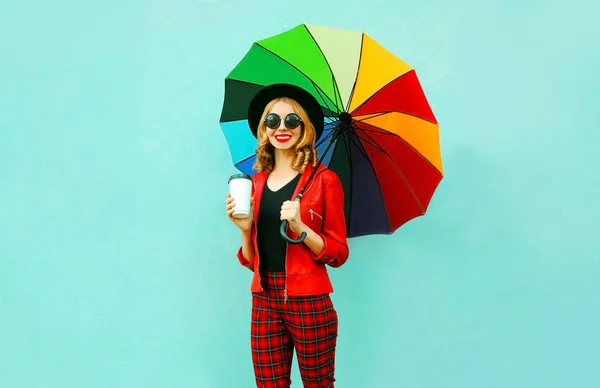 Молодая женщина пьет кофе и держит красочный зонтик ходьба в красной куртке, черная шляпа на фоне синей стены — стоковое фото