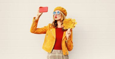 Sonbahar havası! telefondan selfie fotoğrafı çeken kadın gri duvar arka plan üzerinde şehir caddesinde poz fransız bere şapka giyen sarı akçaağaç yaprakları tutan tatlı hava öpücük göndererek kırmızı dudaklar üfleme