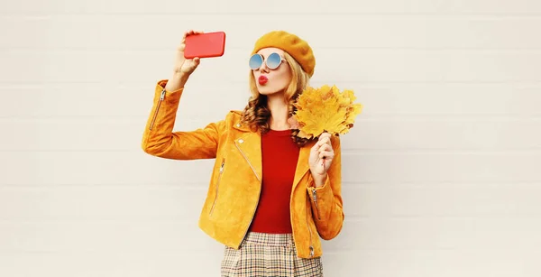가을 무드! 회색 벽 배경을 통해 도시 거리에 포즈를 취하는 프랑스 베레모 모자를 입고 노란색 단풍 잎을 들고 달콤한 공기 키스를 보내는 빨간 입술을 불어 전화로 셀카 사진을 찍는 여성 — 스톡 사진
