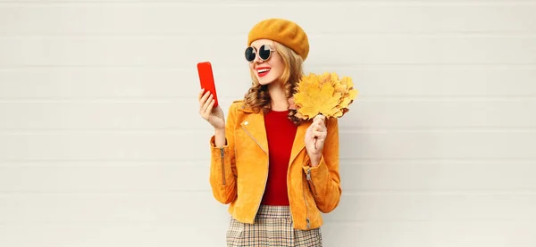 Portrait d'automne femme souriante tenant le téléphone, feuilles d'érable jaune portant béret français sur la rue de la ville sur fond de mur gris — Photo