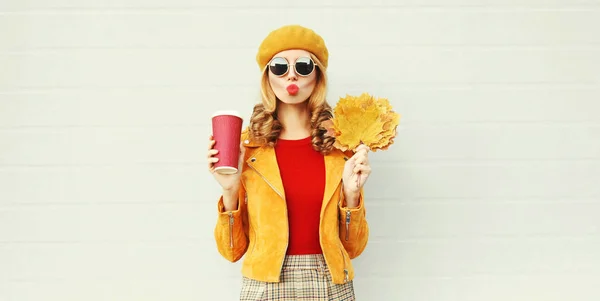 Portrait d'automne femme aux feuilles d'érable jaunes, tasse de café soufflant lèvres rouges envoyant un baiser d'air doux sur fond de mur gris — Photo