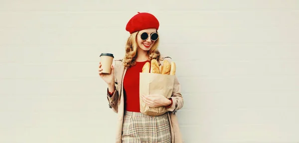 Retrato hermosa mujer sonriente sosteniendo taza de café, bolsa de papel de compras de comestibles con baguette de pan blanco largo sobre fondo de pared gris en blanco — Foto de Stock