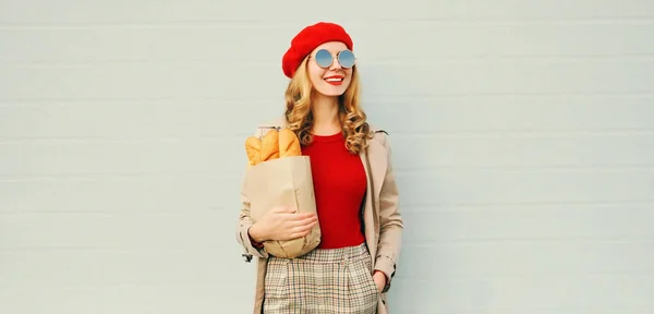 Portrait heureuse jeune femme souriante regardant loin tenant épicerie sac en papier avec longue baguette de pain blanc portant béret rouge sur fond blanc mur gris — Photo