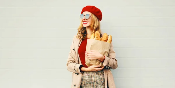 Portrait heureuse jeune femme souriante tenant épicerie sac en papier avec longue baguette de pain blanc regardant loin sur fond blanc mur gris — Photo
