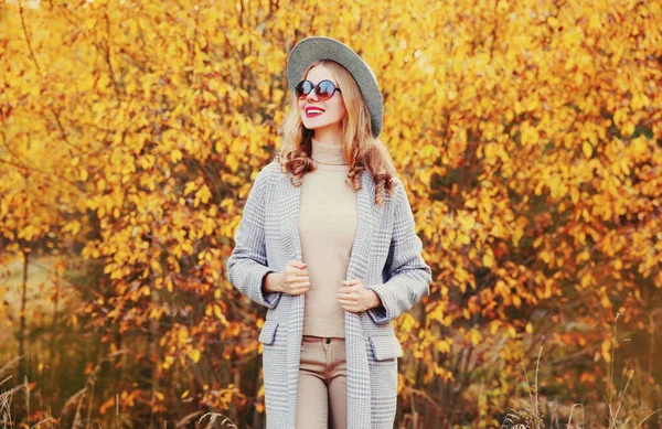 Retrato de otoño elegante mujer sonriente feliz con abrigo gris, sombrero redondo posando sobre el fondo de las hojas amarillas — Foto de Stock