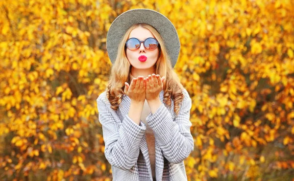 Jesienny portret stylowa kobieta dmuchanie czerwone usta wysyłanie słodkie powietrze pocałunek noszenie szary płaszcz, okrągły kapelusz na żółte liście tło — Zdjęcie stockowe