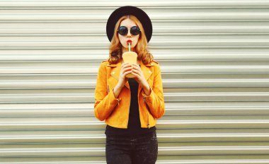 Şık bir kadın meyve suyu içiyor sarı ceketli, siyah yuvarlak. 