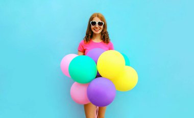 Bir sürü balonu olan mutlu, genç bir kadın mavi arka planda şort ve pembe tişört giyerek eğleniyor.