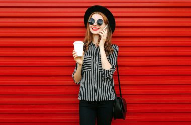 Genç bir kadın elinde kahve fincanı, elinde siyah yuvarlak bir şapka ve kırmızı duvar arkasında el çantası olan akıllı telefondan arıyor.