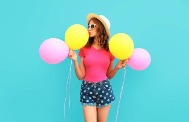 Yazın, sarı pembe balonları öpen, mavi arka planda şort ve hasır şapka takan hoş bir genç kadının renkli görüntüsü.