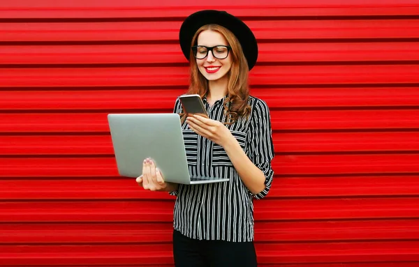 在红墙背景下拿着笔记本电脑看手机的现代女性 — 图库照片