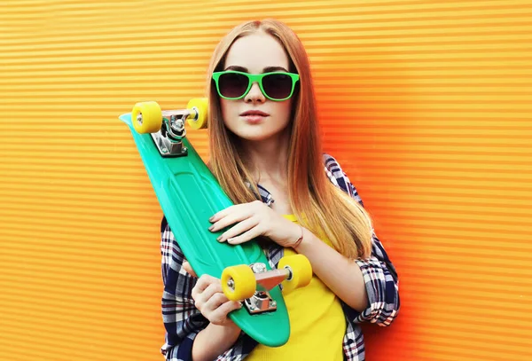 肖像若いですブロンド女性とともに緑のスケートボードを身に着けていますサングラス以上カラフルなオレンジ色の壁の背景 — ストック写真