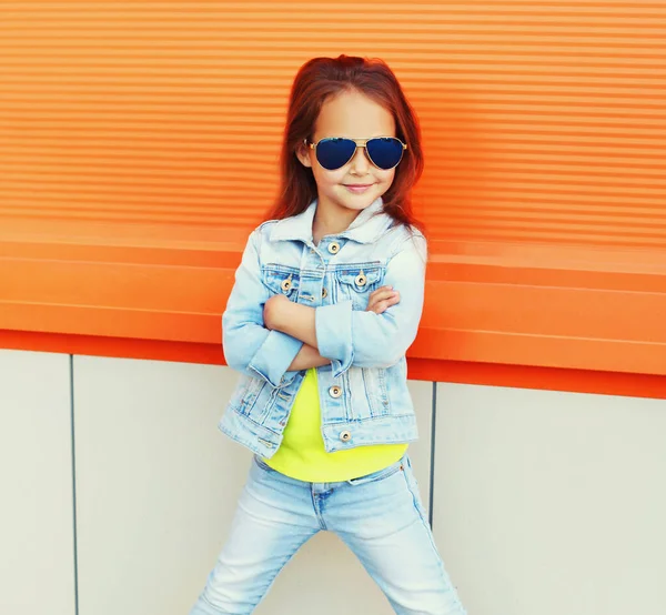 照片上的小女孩戴着太阳镜 穿着牛仔裤夹克 背景是橙色的 — 图库照片