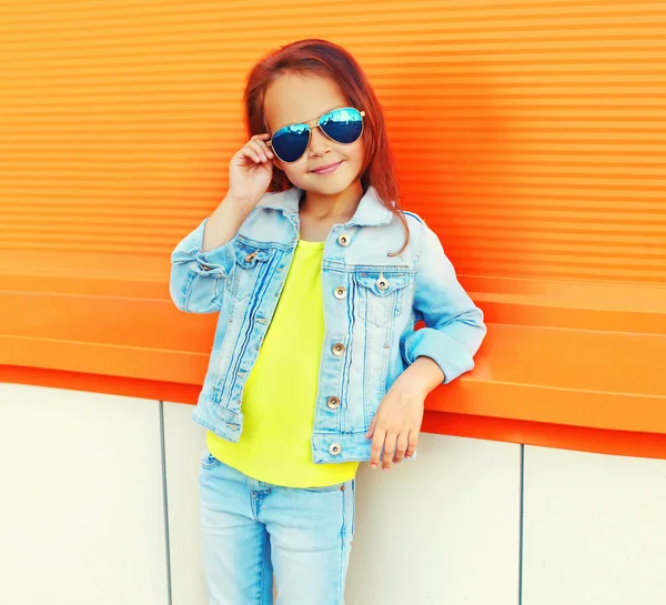 照片上的小女孩戴着太阳镜 穿着牛仔裤夹克 背景是橙色的 — 图库照片