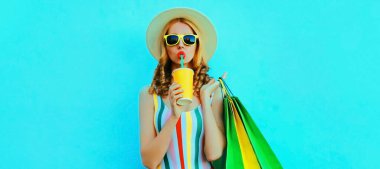 Renkli tişörtlü alışveriş torbalarıyla meyve suyu içen genç bir kadının portresi, mavi arka planda yaz tatili şapkası.
