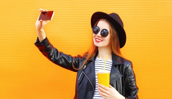 快乐地微笑着的年轻女子通过电话在橙色墙上的背景上放着一杯果汁 给自己拍照 — 图库照片
