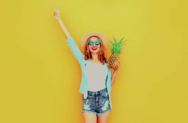 Yaz görüntüsü mutlu kadın ananasla elini kaldırıyor hasır şapka takarak eğleniyor, şortu sarı arka planda.