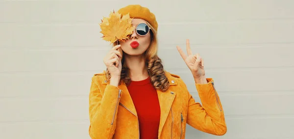 秋のポートレートの魅力的な女性とともに黄色のカエデの葉 女性モデル吹いて赤い唇送信甘いです空気キス身に着けているフランス語ベレー帽上の灰色の背景 — ストック写真
