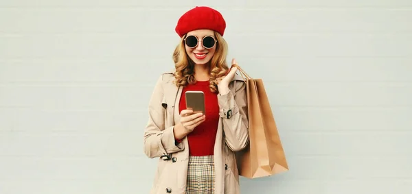 スマートフォンを持つ笑顔の女性の秋の肖像画とショッピングバッグコートを着て グレーの背景に赤いフランス語ベレー帽 — ストック写真