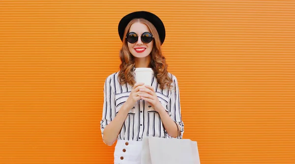 一个迷人的笑容可亲的女人 戴着咖啡杯 头戴黑色圆帽 背景是橙色的条纹白衬衫的购物袋 — 图库照片