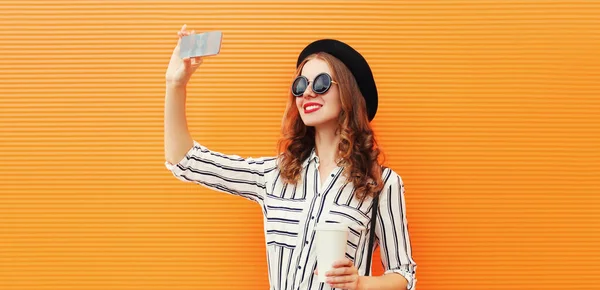 白いストライプのシャツを着たコーヒーカップ オレンジの背景に黒い丸帽子を身に着けているスマートフォンで写真を撮るスタイリッシュな若い笑顔の女性 — ストック写真