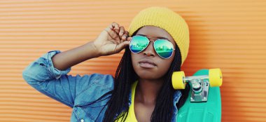 Kaykaylı, güneş gözlüğü takan, portresi turuncu duvarlı, şapkalı genç bir Afrikalı kadın portresi.