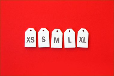 Kırmızı arka planda beyaz ahşap etiketlerde XS, S, M, L, XL giyim ölçüleri.