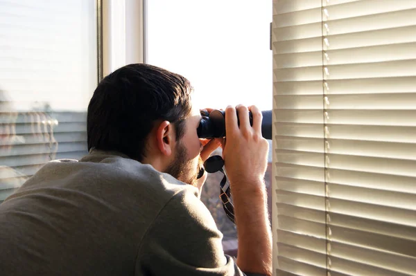 Young man standing looking through a glass window with binoculars as he watches something in the distance.