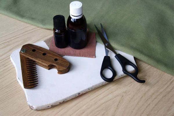 Male wooden comb for beard and hair, care oils and scissors for haircuts on white desk. Compact folding comb that you can always take with you. Beard and hair care concept.