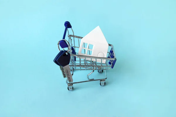 Mini shopping trolley with a white house and keys on a blue background. The concept of buying or renting a house, apartment, cottage and more.