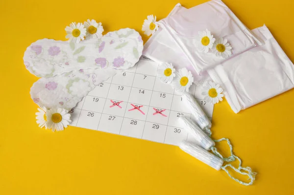 Menstrual pads and tampons on menstruation period calendar with chamomiles on yellow background. The concept of female health, personal hygiene during critical days.