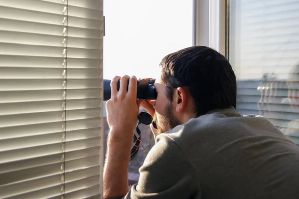 Young man standing looking through a glass window with binoculars as he watches something in the distance.