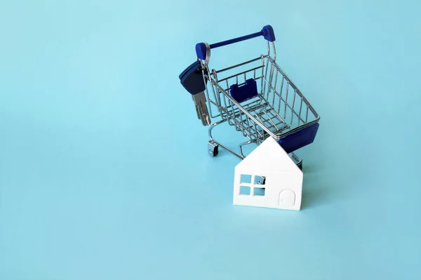 Mini shopping trolley with a white house and keys on a blue background. The concept of buying or renting a house, apartment, cottage and more.