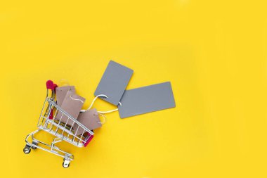 Küçük el çantaları ve sarı arka planda boş gri etiketleri olan mini alışveriş arabası. Mağazalarda ve internette alışveriş kavramı.