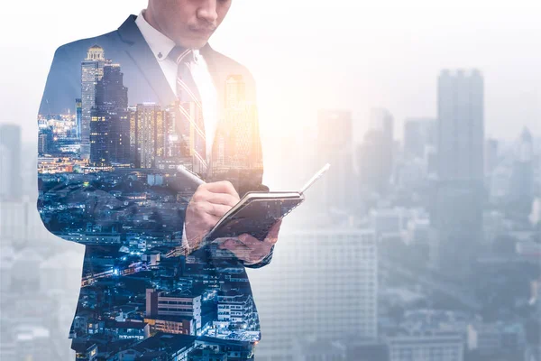 The double exposure image of the businessman standing and writing on his notebook during sunrise overlay with cityscape image. The concept of management, business, city life and professional.