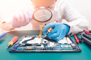 Asyalı teknisyen, laboratuvarda tabletin ana kartını tamir etmek için büyüteç kullanıyor. bilgisayar donanımı, cep telefonu, elektronik, onarım ve teknoloji kavramı.