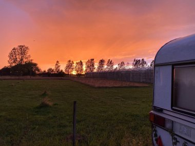 Büyük portakal rengi günbatımı ve karavanın huzuru, karavanın karavanı, ağaçlar ve çimenlerle dolu yeşillik.