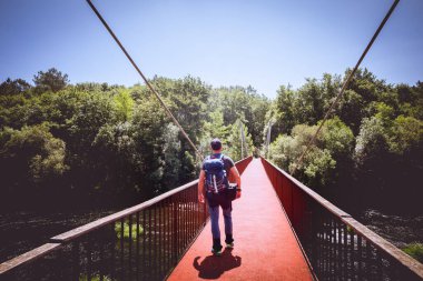 Sırt çantalı ve şapkalı bir adam nehrin ortasındaki bir köprüden geçiyor.