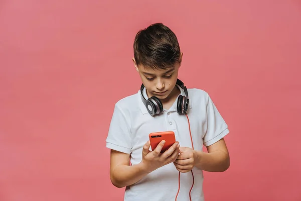 Beyaz tişörtlü ve kulaklıklı yakışıklı bir çocuk, akıllı telefonunu tutuyor, internette yeni müzikler arıyor, pembe stüdyo arkaplanı üzerinden. Gençlik ve çağdaş insanlar kavramı.