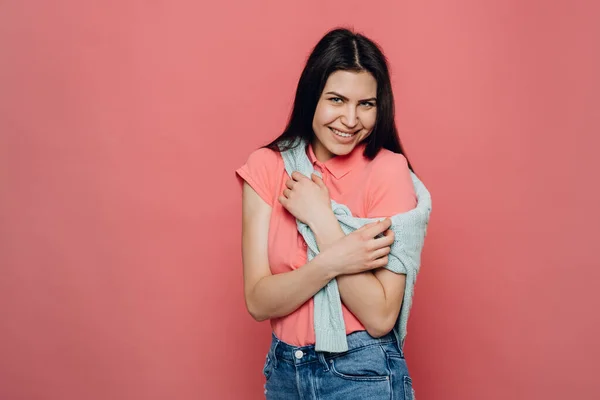 Genç, güzel esmer kadın, günlük tişört giyiyor, pembe arka planda süveter giyiyor utangaç ve neşeli bir kadın gülümsüyor, neşeli bir ruh hali içinde kendine sarılıyor. Samimi insan duyguları..