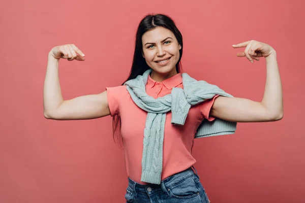 Young cute brunette dressed in pink t-shirt and turquoise sweater tied up over her shoulders, smiles happily, points at herself, satisfied by her physical state, stands over pink background.