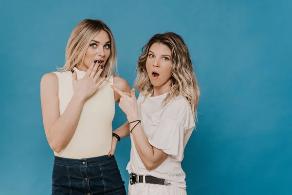 Two blondes in casual clothes gossip, one in shock from the story of her friend looks in amazement at the camera. Girls over blue background. People emotions.