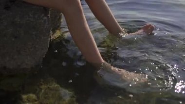 Büyük bir kayanın üzerinde oturan ve ayaklarını suya sokan bir gencin portresi. Çocuk elinde bir dalış maskesi tutuyor. Deniz, sabah, ağır çekim.