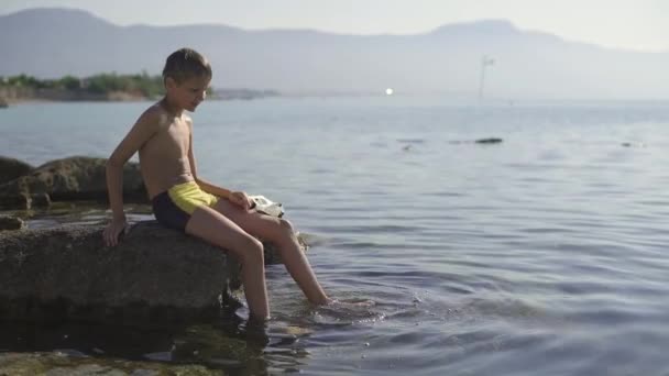 Підліток сидить на великій скелі і вимочує ноги у воді. Хлопець тримає маску для дайвінгу в руці. Море, ранок, повільний рух . — стокове відео