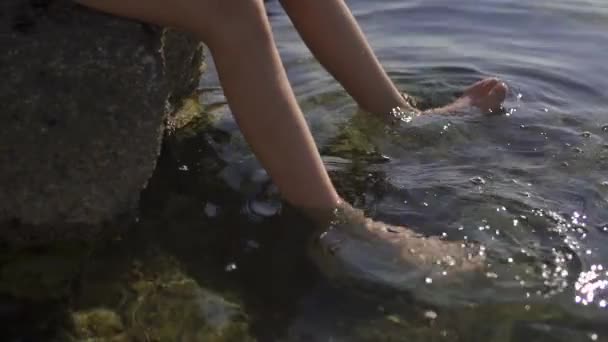 Portret van een tiener die op een grote rots zit en zijn voeten in het water doordrenkt. De jongen houdt een duikmasker in zijn hand. Zee, ochtend, slow motion. — Stockvideo