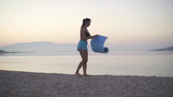 A beautiful girl spreads a karemat by the sea and begins to train. Sports, yoga, beach. — Stock Video