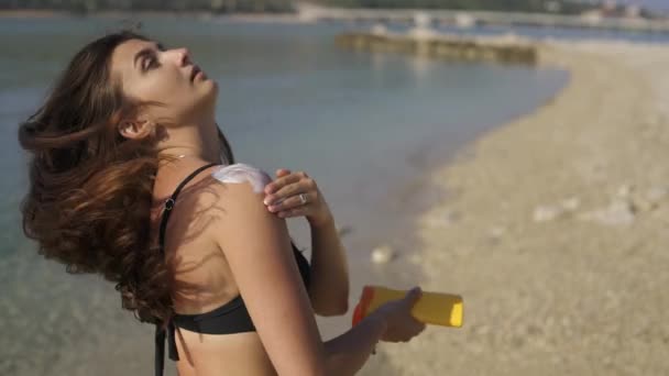 Beautiful girl smears herself with sunscreen standing on the beach. Morning on a deserted beach. — Stock Video