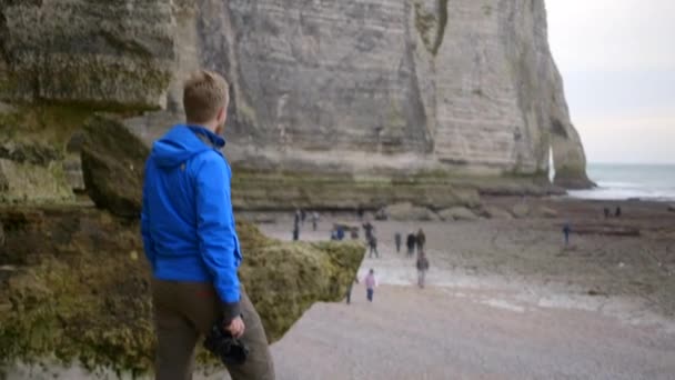 Un fotógrafo profesional toma fotos de su próxima toma. La playa de Etretat. Cámara profesional. Pasos sobre roca escarpada. Disparando a mano. Etretat Francia — Vídeos de Stock