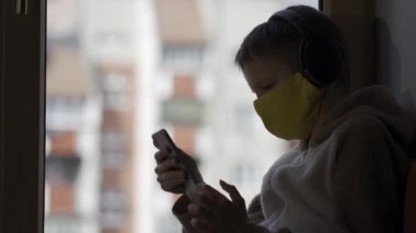Koruyucu maskeli çocuk bir pencere pervazında oturuyor ve elinde telefon var. Karatnin.