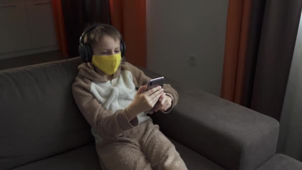 De man met het beschermend masker zit op de bank naar muziek te luisteren in een koptelefoon. Quarantaine 2020 — Stockvideo